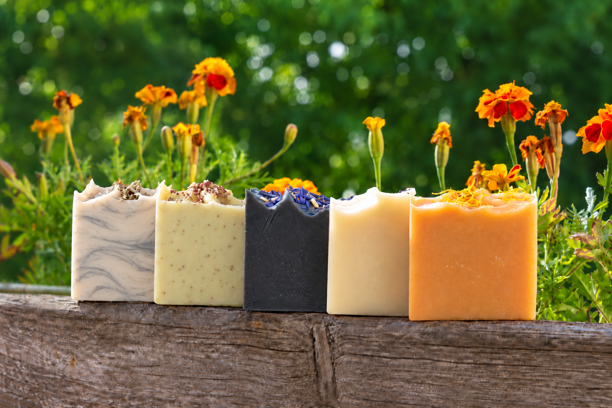 Fünf handgemachte Naturseifen auf einem Holzbalken vor blühenden Gartenblumen.